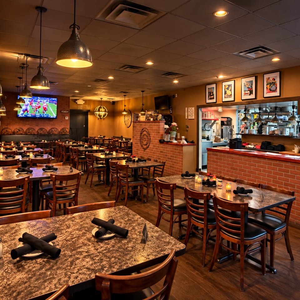 Warmly lit restaurant dining room with set tables, pendant lights, brick bar, framed artwork, and televisions showing sports.