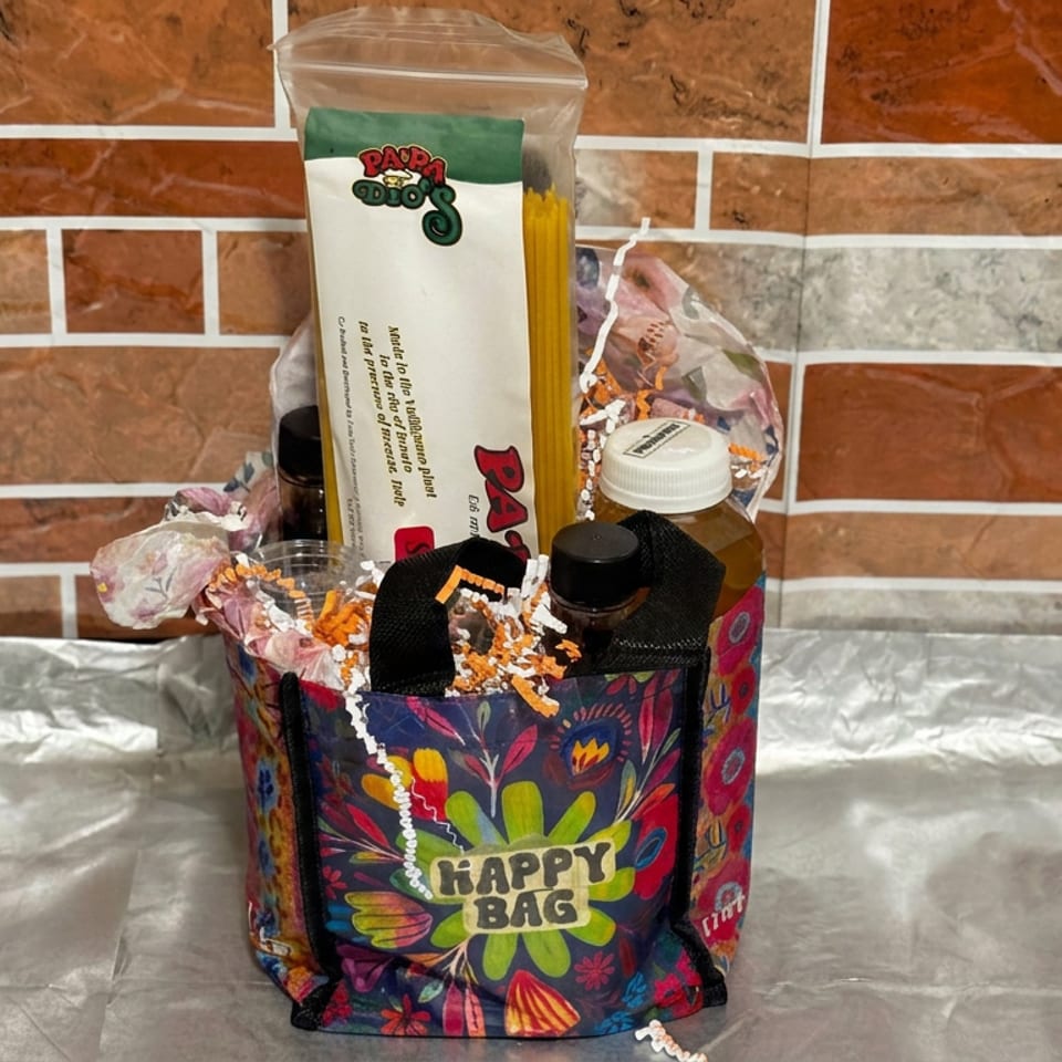 Colorful floral “Happy Bag” gift basket filled with treats, a card, bottle, and decorative paper against a brick wall.