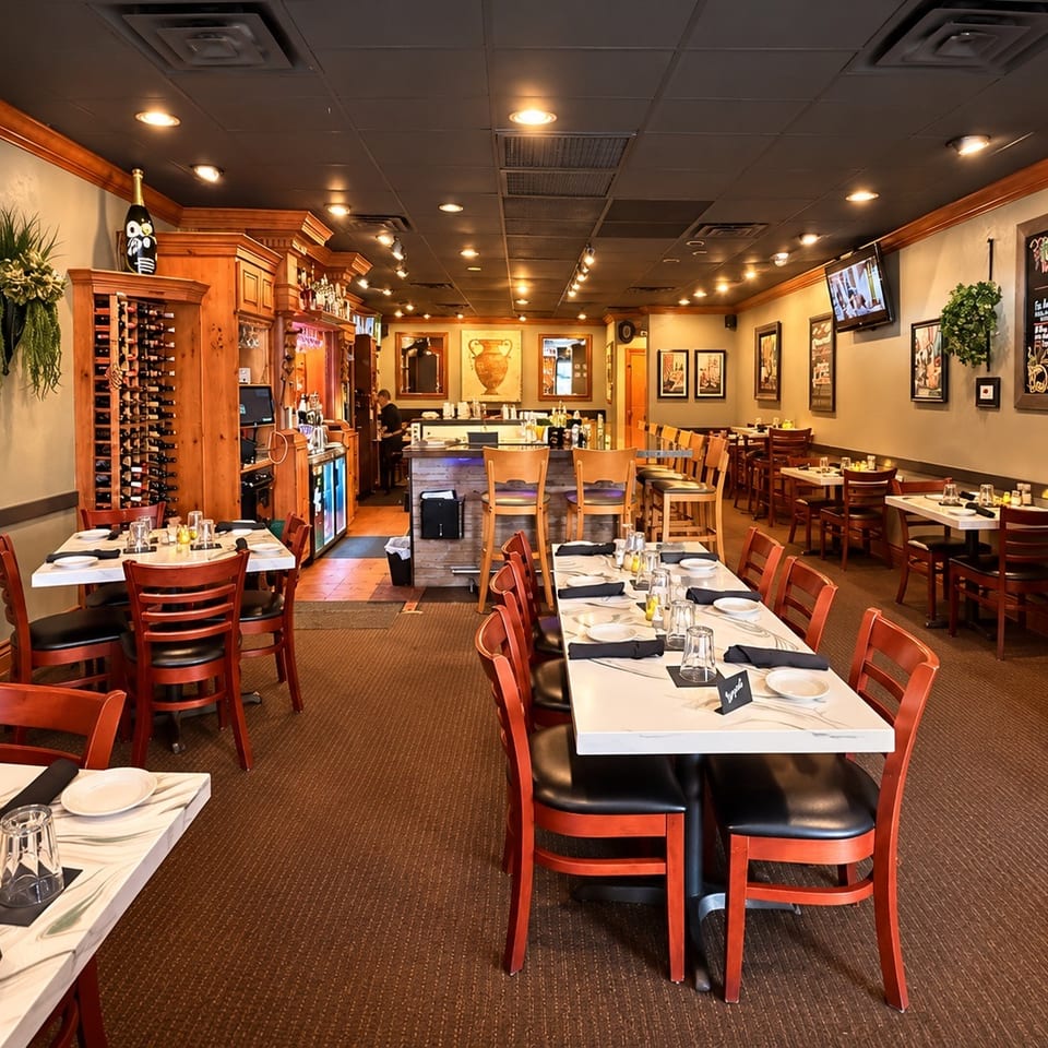 Warmly lit restaurant dining room with set tables, wooden chairs, a bar, wine rack, artwork, and chalkboard menu.